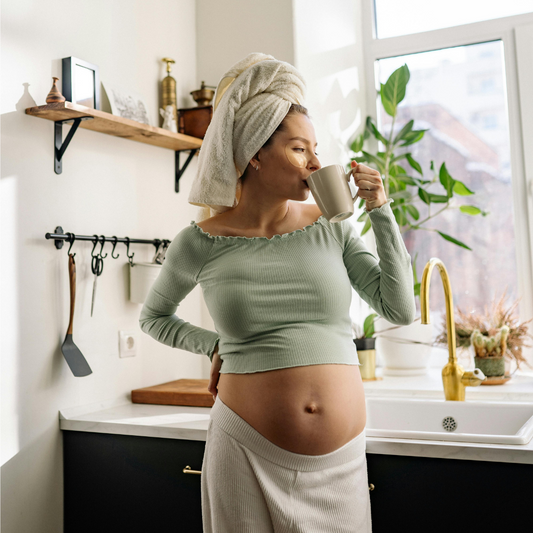 The image shows a pregnant woman standing in a bright kitchen, holding a hot drink. She is surrounded by greenery visible through the windows, and her expression and posture exude a healthy, glowing appearance.