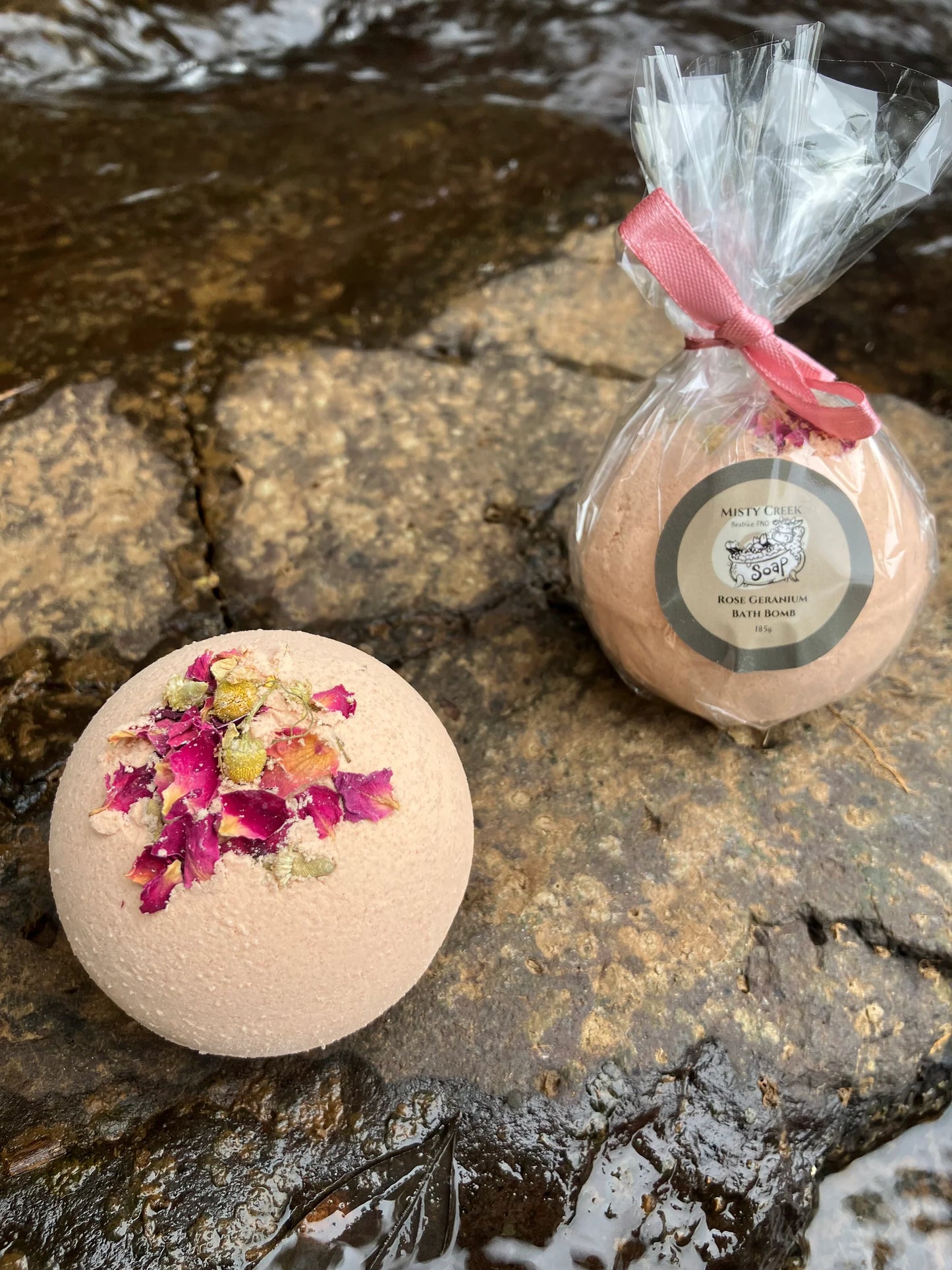 Pink Rose Geranium bath bomb placed on a rock at a waterfall, surrounded by delicate rose petals and a sprig of greenery, exuding a luxurious and calming vibe.