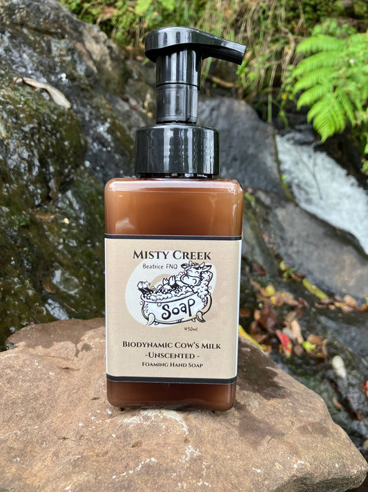 A bottle of Biodynamic Cow’s Milk Foaming Unscented Liquid Hand Soap resting on a smooth rock near a cascading waterfall, surrounded by lush greenery, highlighting its natural and eco-friendly essence.