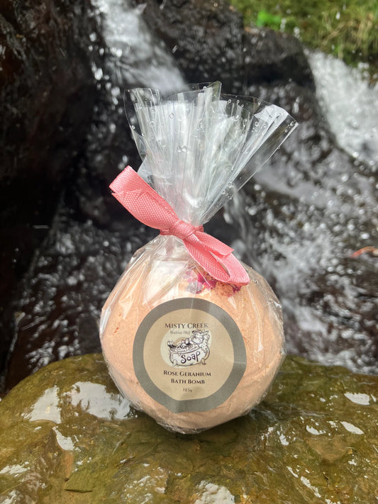 Pink Rose Geranium bath bomb placed on a rock at a waterfall, surrounded by delicate rose petals and a sprig of greenery, exuding a luxurious and calming vibe.